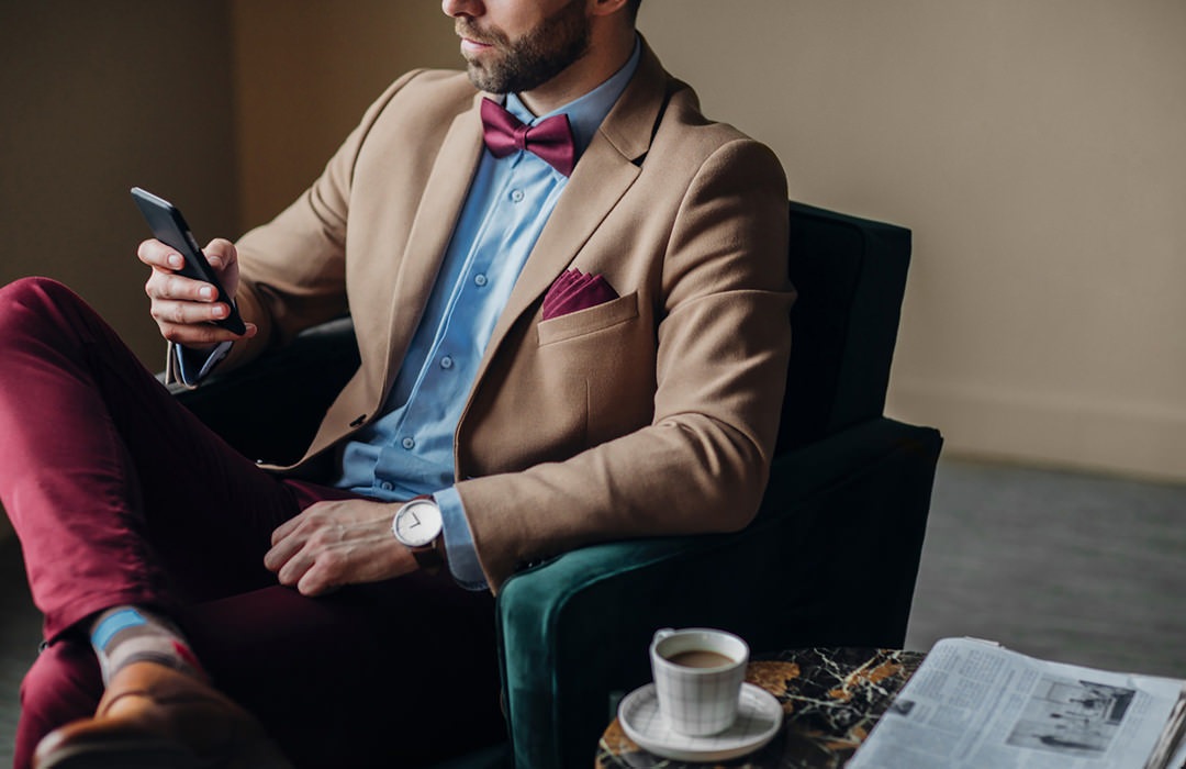 dapper young man sits in lounge chair and looks at cell phone with a cup of coffee at his side